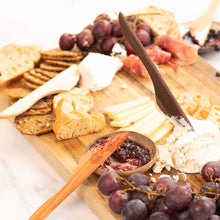 Three wooden spreaders on a cutting board with cheese, crackers, spreads, and grapes.