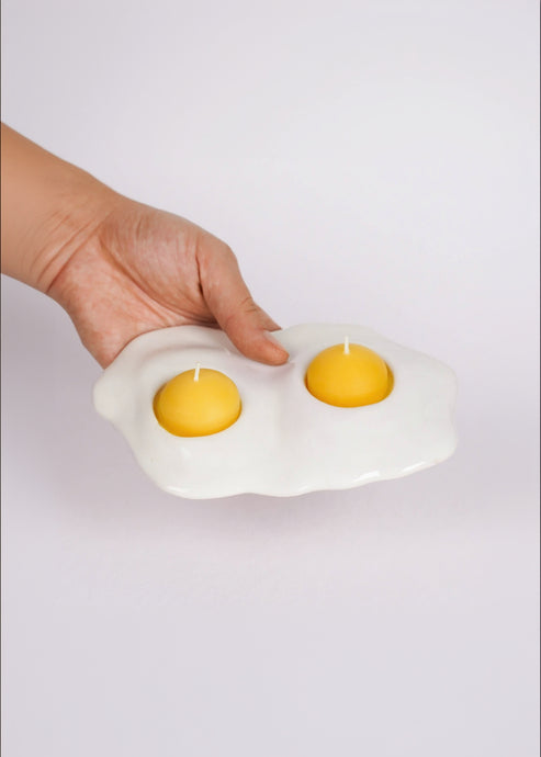 Hand holding a white egg-shaped candle holder with two yellow candles inside on a light gray background