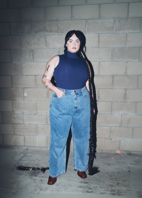 Person wearing a blue sleeveless top and jeans standing against a brick wall.